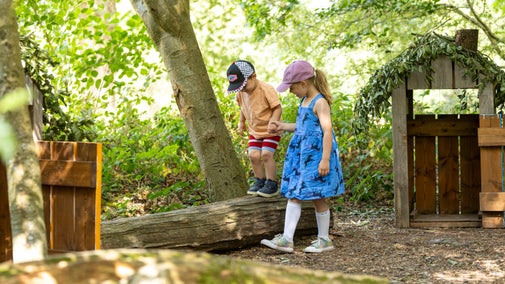 A young girl helps a little boy balancing on a log at Packwood House, Warwickshire